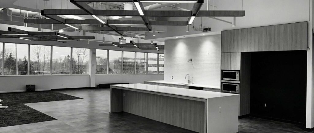 Modern open-concept office kitchen with large island, geometric ceiling lights, and full wall of windows.