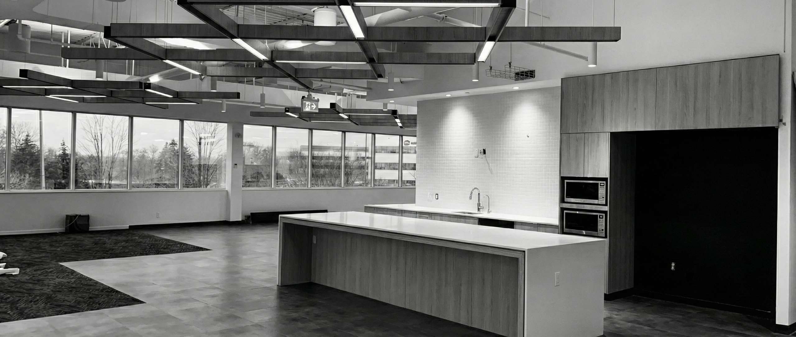 Modern open-concept office kitchen with large island, geometric ceiling lights, and full wall of windows.