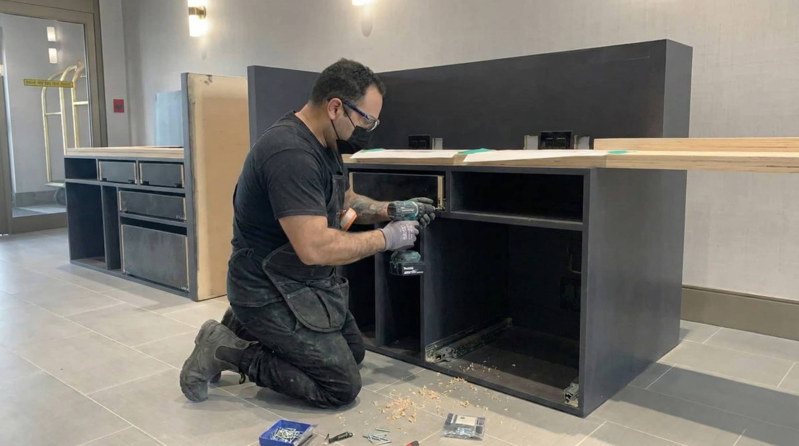 Masood installing cabinets in modern office workspace using drill and safety gear.